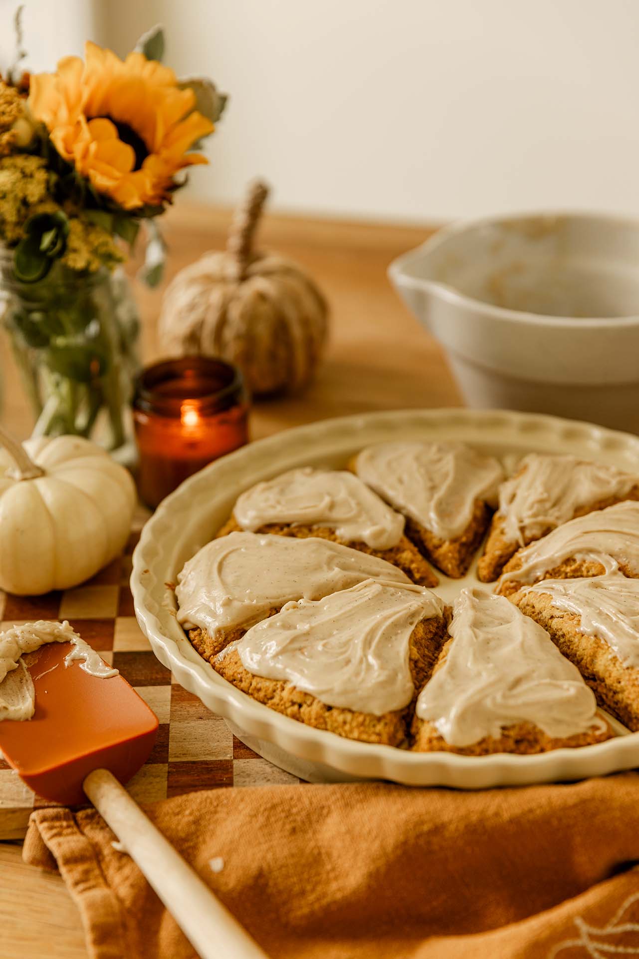 Freshly baked pumpkin scones with brown butter glaze arranged in a cream pie dish on a wooden table. The golden scones are swirled with vanilla-speckled glaze, surrounded by a lit amber candle, white pumpkin, and a sunflower bouquet — a warm, inviting autumn baking scene.